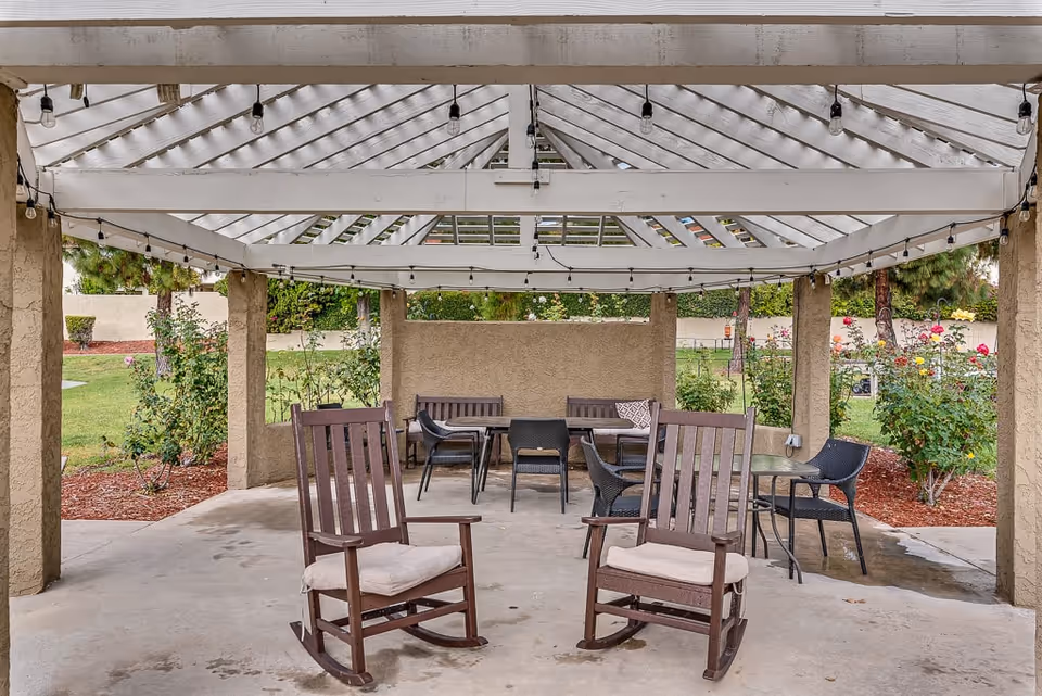 Outdoor covered seating area with a white pergola roof, two wooden rocking chairs with cushions in the foreground, and several tables and chairs in the background surrounded by garden landscaping with bushes and flowers.