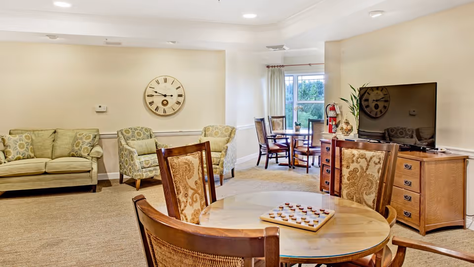 Communal living room with upholstered sofas and chairs, round tables and a TV on a wooden cabinet.