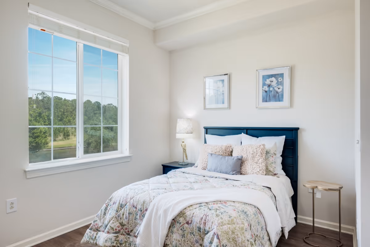 A bright bedroom with a large window showing a view of trees outside. The room features a bed with a dark blue headboard, floral bedding, multiple pillows, a bedside table with a lamp, and two framed floral artworks on the wall above the bed.