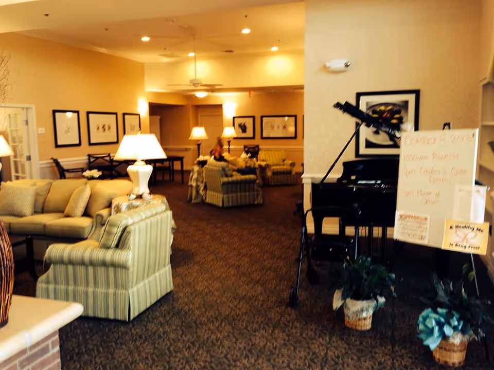 A cozy living room area in Southwest Assisted Living featuring several upholstered armchairs and sofas arranged around lamps and tables. The room has warm lighting, framed artwork on the walls, a piano with a music stand, and a whiteboard with event information. There are also some potted plants on the floor near the piano.
