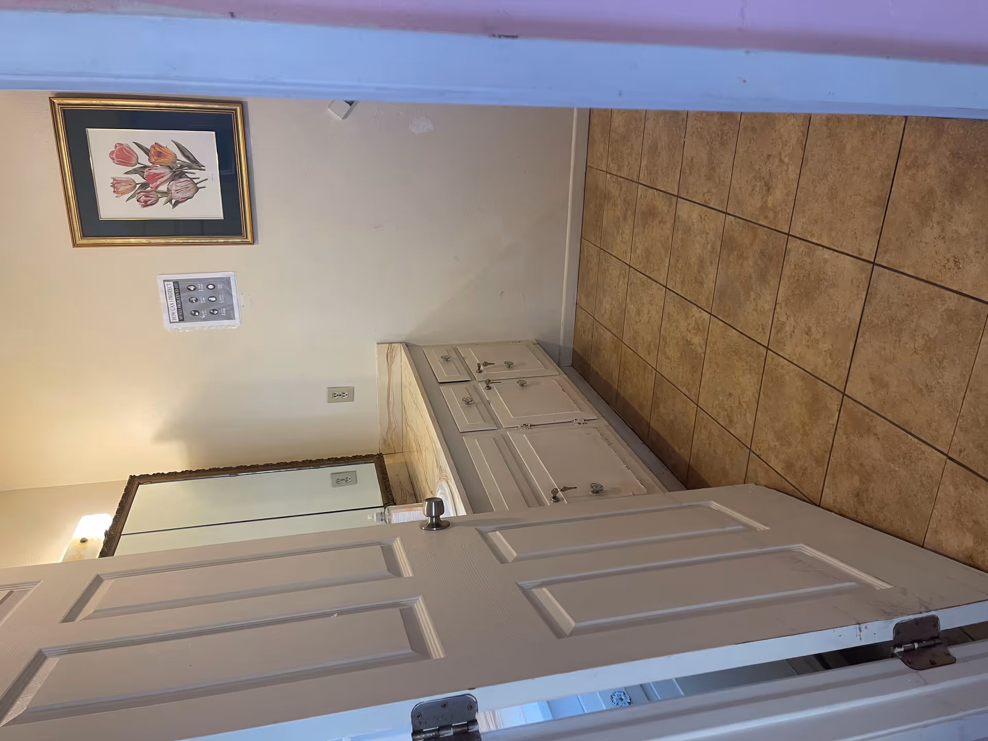 A bathroom with a tiled floor, a white door partially open, a countertop with a sink and a large mirror above it. There is a framed picture of pink and orange tulips on the wall and a small poster with COVID-19 protection tips.