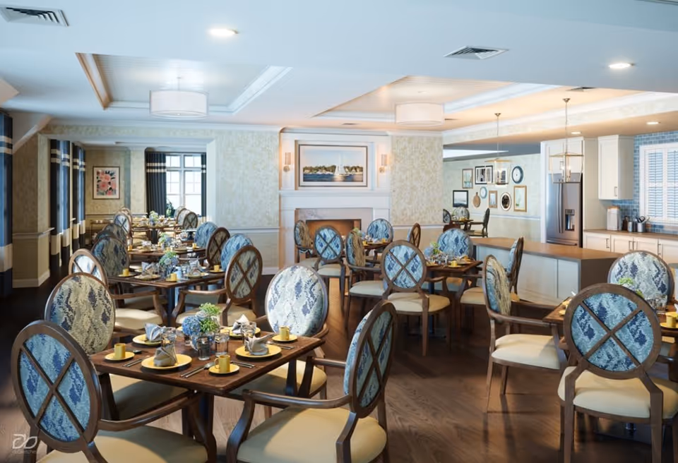 A bright and elegant dining room with multiple wooden tables and chairs featuring blue patterned upholstery. Each table is set with plates, cups, napkins, and silverware. The room has a cozy fireplace with a painting above it, large windows with curtains, and a kitchen area with white cabinets and stainless steel refrigerator in the background.