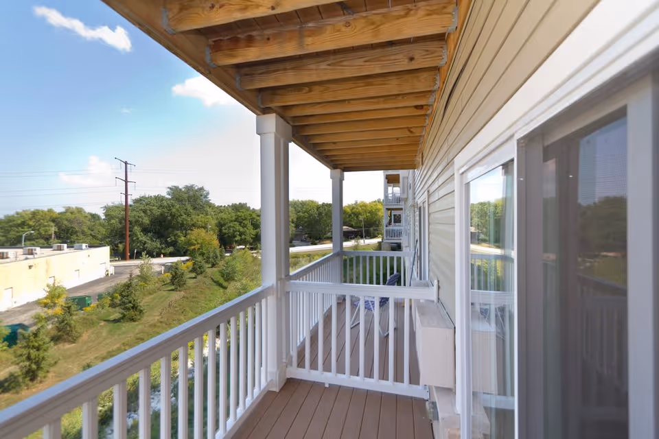 Covered apartment balcony with white railing and wooden ceiling overlooking trees and nearby buildings.