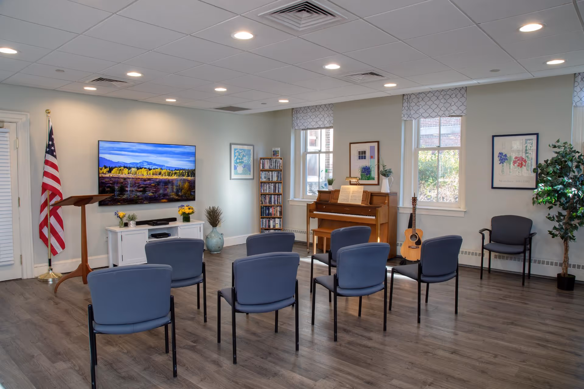 A well-lit common room with six blue chairs arranged in two rows facing a television mounted on the wall. The room features a wooden piano with sheet music, a guitar resting against the wall, framed artwork, an American flag, a small podium, and a potted plant. Large windows with patterned valances allow natural light to enter.