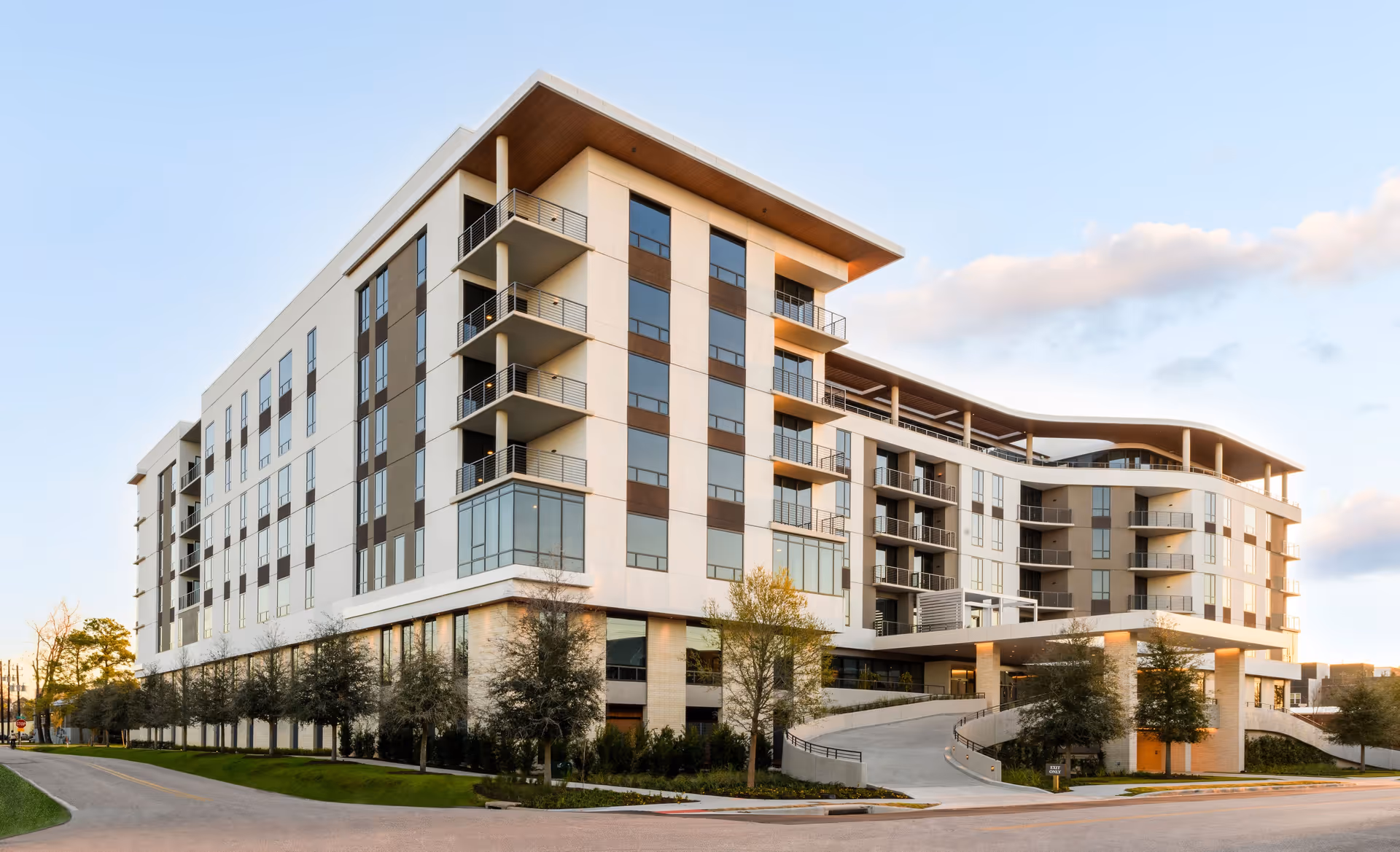 Modern multi-story senior living facility building with large windows and balconies, surrounded by trees and a driveway, under a partly cloudy sky during daylight.