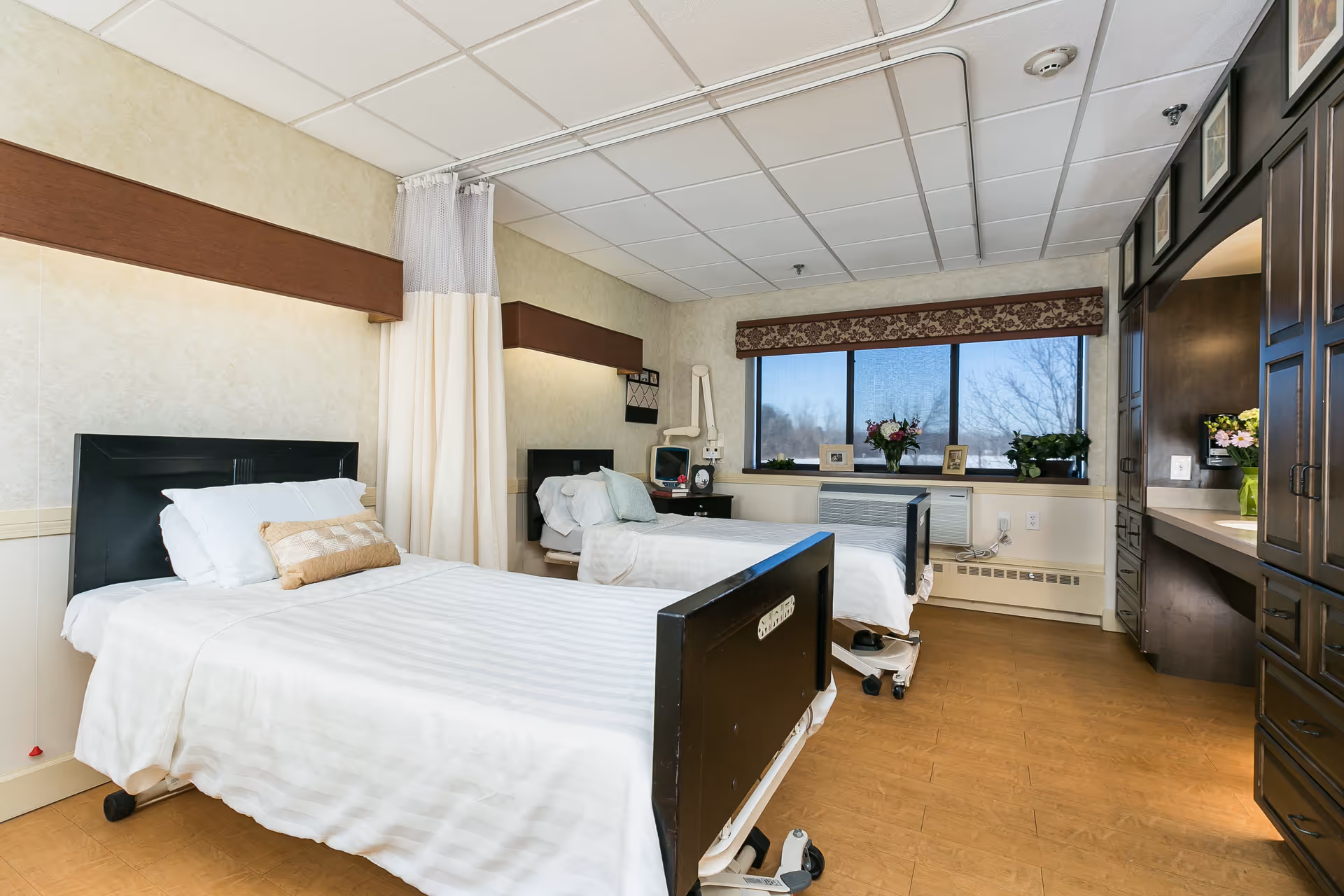 A bright nursing facility bedroom with two hospital beds separated by a privacy curtain, a large window, and built-in cabinetry.