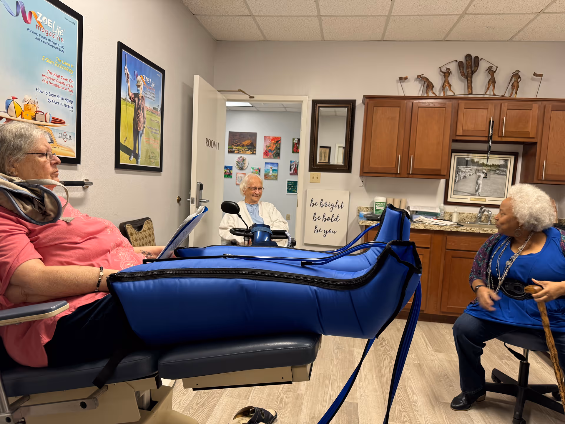 Three elderly women in a room at Glencroft Center for Modern Aging. One woman is seated on a medical recliner with blue compression leg sleeves, another woman is sitting on a chair holding a cane, and a third woman is in a wheelchair near an open door labeled 'Room 1'. The room has wooden cabinets, a granite countertop, framed pictures on the walls, and a decorative metal sculpture on top of the cabinets.