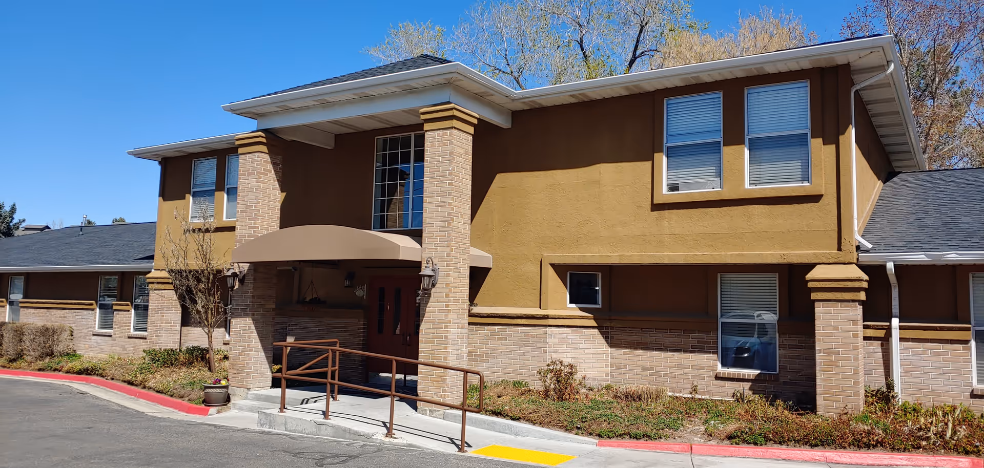 Front entrance of a two-story tan brick and stucco assisted living building with a covered entry, wheelchair ramp, and landscaping.