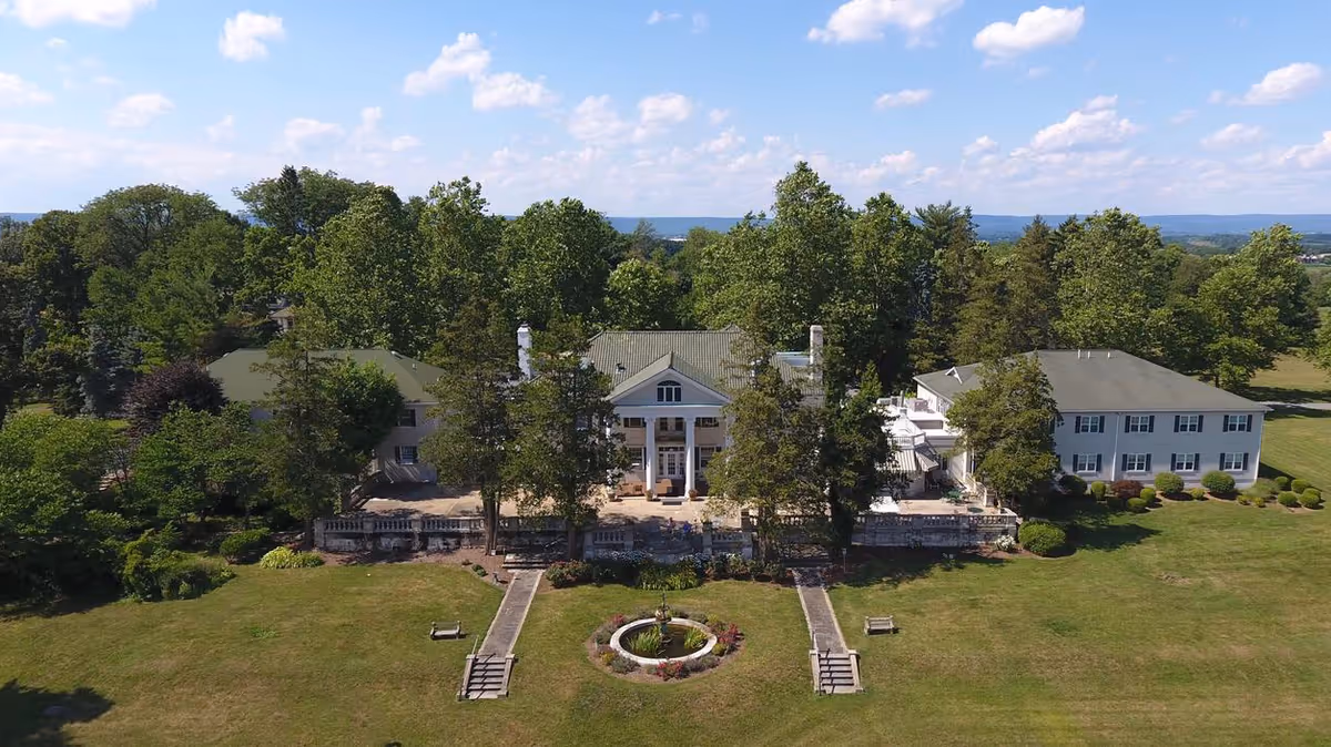 Aerial view of a large estate building surrounded by trees and greenery under a partly cloudy sky. The building has a central section with tall white columns and two wings extending on either side. In front of the building is a circular garden feature with a fountain and two benches on a grassy lawn.