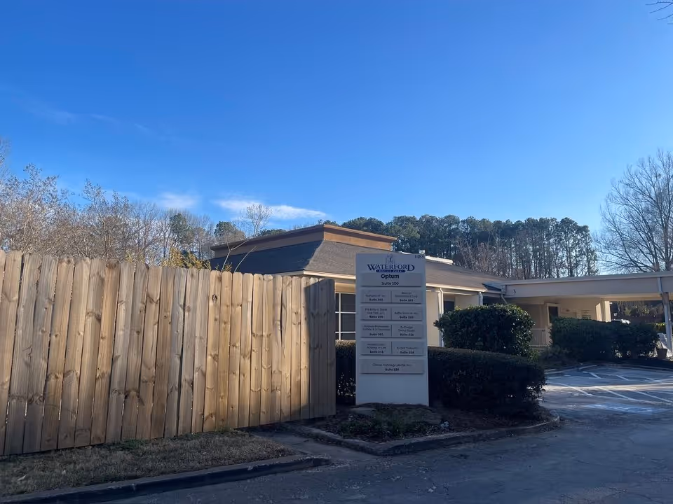Front exterior of a single-story senior living building with a wooden fence, landscaping, and a directory sign under a clear blue sky.