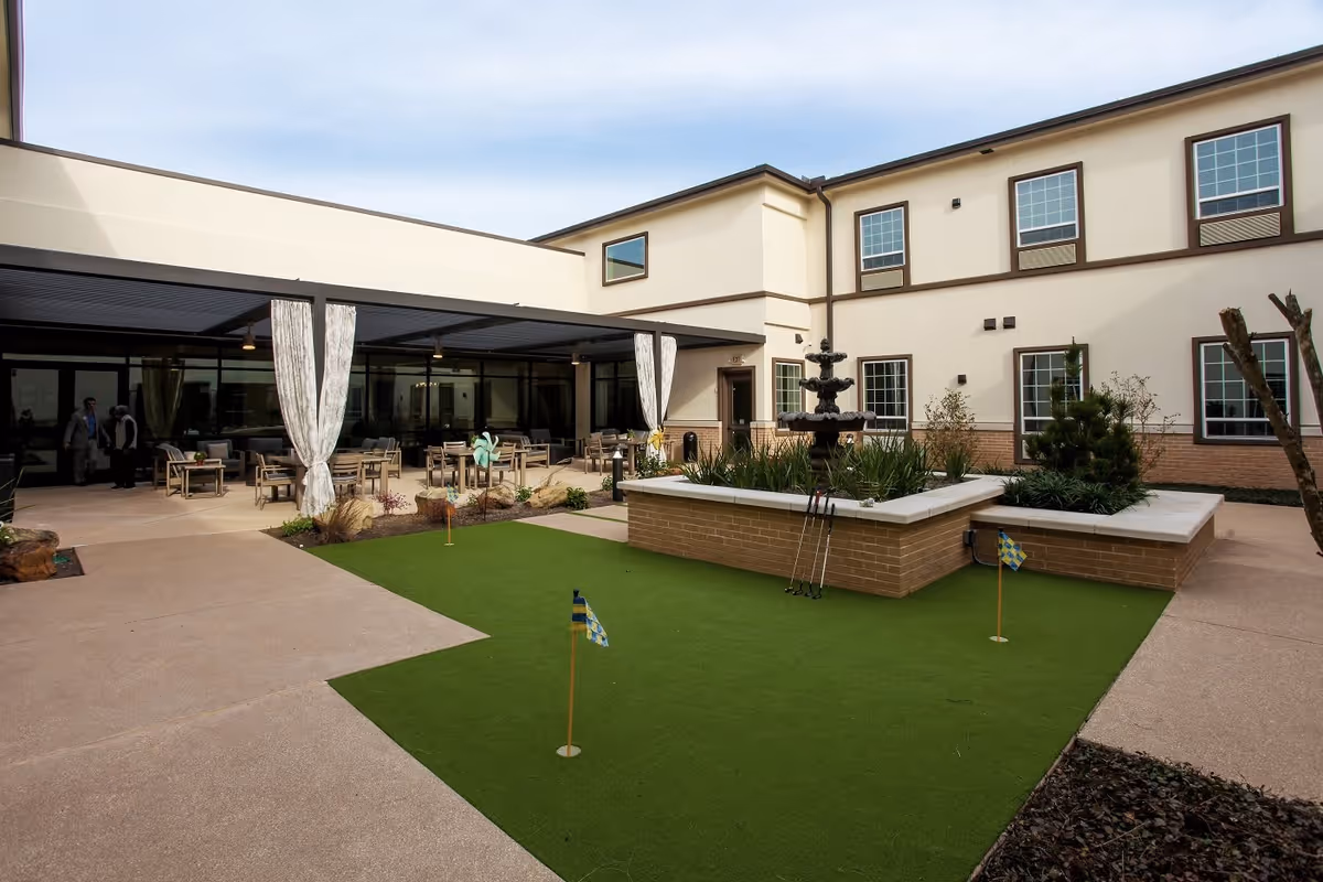 Outdoor courtyard area at The Legacy at Long Meadow featuring a small putting green with flags, a central raised planter with a multi-tiered fountain, and a covered patio with seating and tables. The building surrounds the courtyard with multiple windows visible.