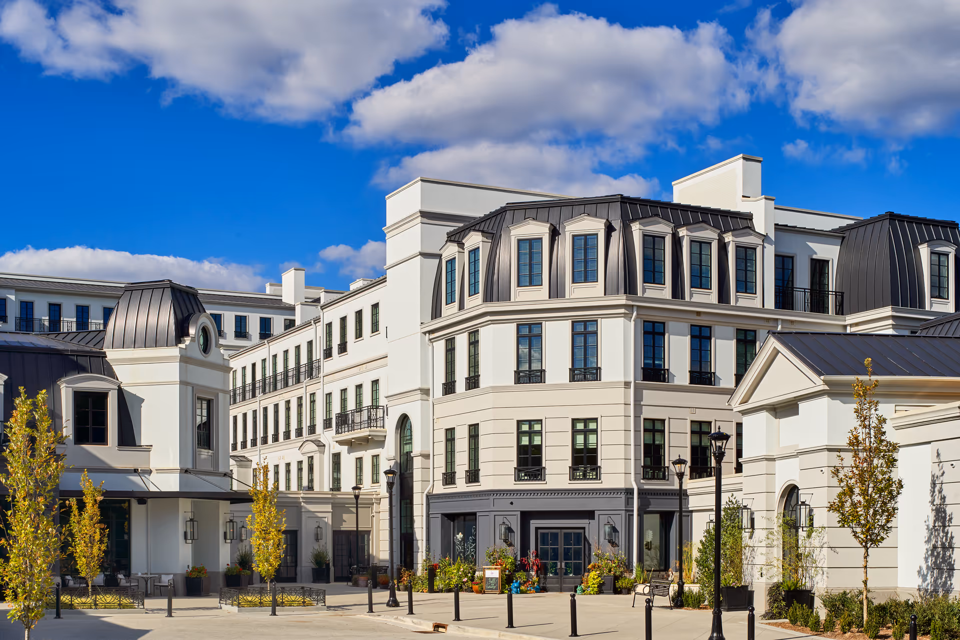Exterior view of a large, elegant senior living facility building with white walls, multiple windows, and black metal roofing under a blue sky with scattered clouds. There are small trees and street lamps in front of the building.