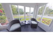 A small enclosed sunroom with two wicker armchairs and a round side table facing large windows that overlook landscaped grounds and a road.