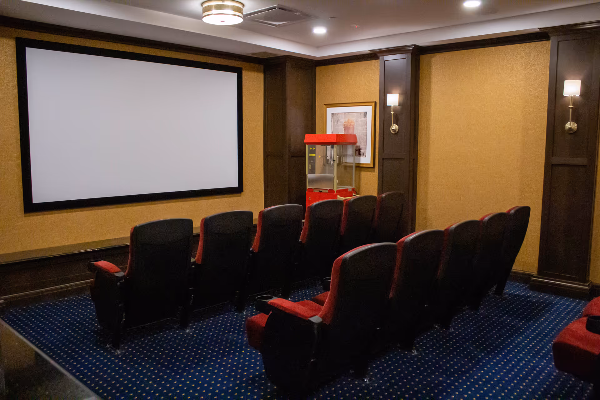 A small movie theater room with two rows of red and black cushioned seats facing a large blank projection screen. There is a red popcorn machine in the corner and framed artwork on the wall. The room has dark wood paneling, beige walls, blue carpet with a dotted pattern, and wall-mounted lights.