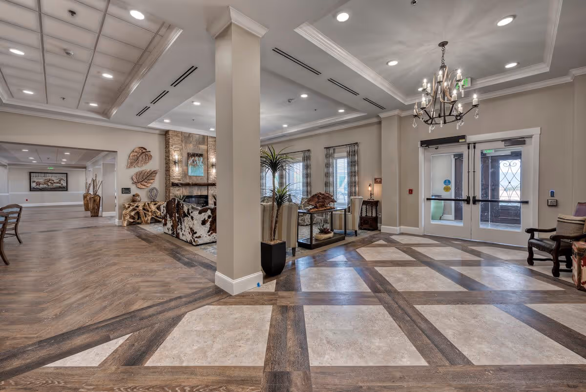 Spacious and well-lit senior living facility lobby with a patterned floor combining wood and tile, a chandelier hanging from the ceiling, double glass doors leading outside, and a cozy seating area with armchairs and a stone fireplace. Decorative elements include large leaf wall art and potted plants.