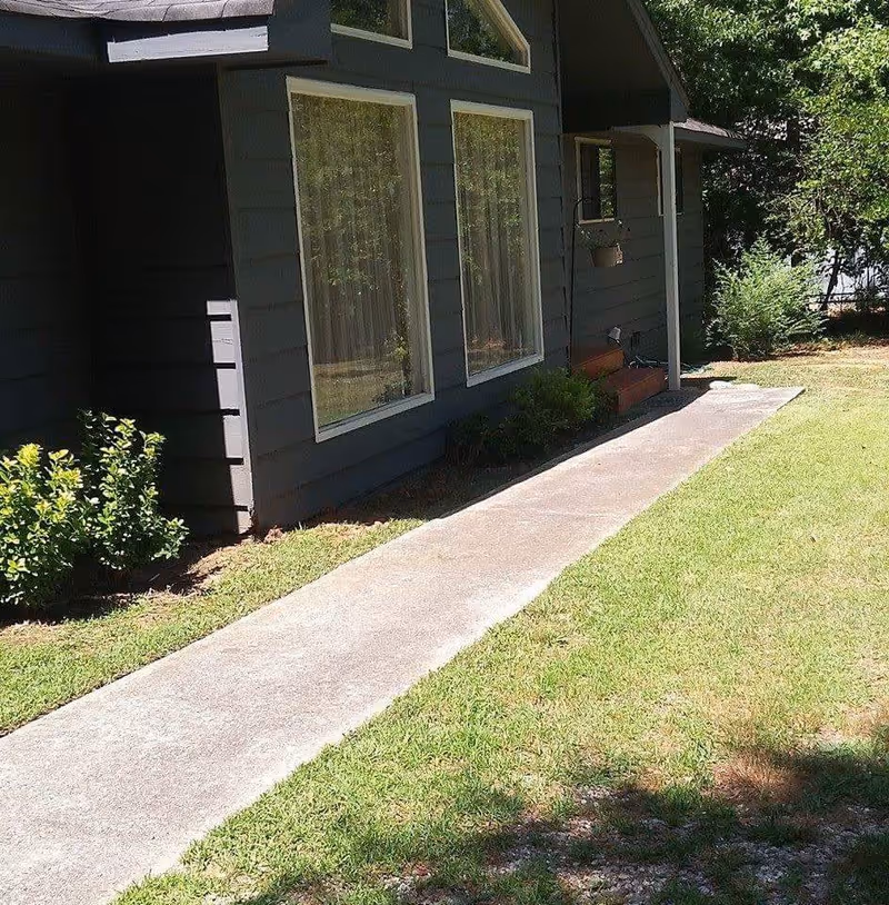 Side view of a dark gray building with two large windows and a small covered porch with steps, surrounded by green grass and bushes.