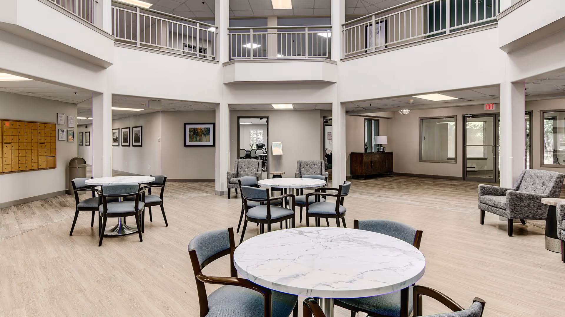 Spacious and bright common area in a senior living facility with round marble-top tables and chairs arranged for seating. The room features light wood flooring, white walls, and a high ceiling with a second-floor balcony overlooking the area. There are framed pictures on the walls, a set of mailboxes on the left side, and comfortable armchairs placed around the space.