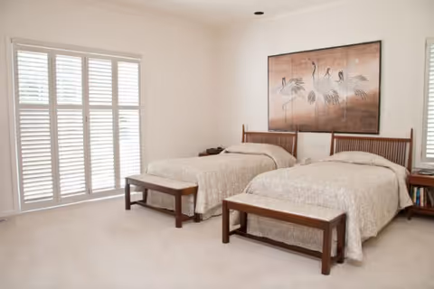 A bright bedroom with two twin beds, each with beige bedspreads and wooden headboards. At the foot of each bed is a wooden bench. The room has light-colored walls and carpet, with large windows covered by white plantation shutters. Above the beds is a large framed artwork featuring birds.