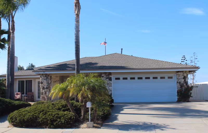 Single-story home front with a two-car garage, stone accents, palm trees, driveway and an American flag on the roof.