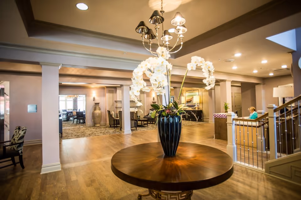 Spacious senior living facility lobby with a round table holding a large vase of white orchids, seating areas, and a reception desk.
