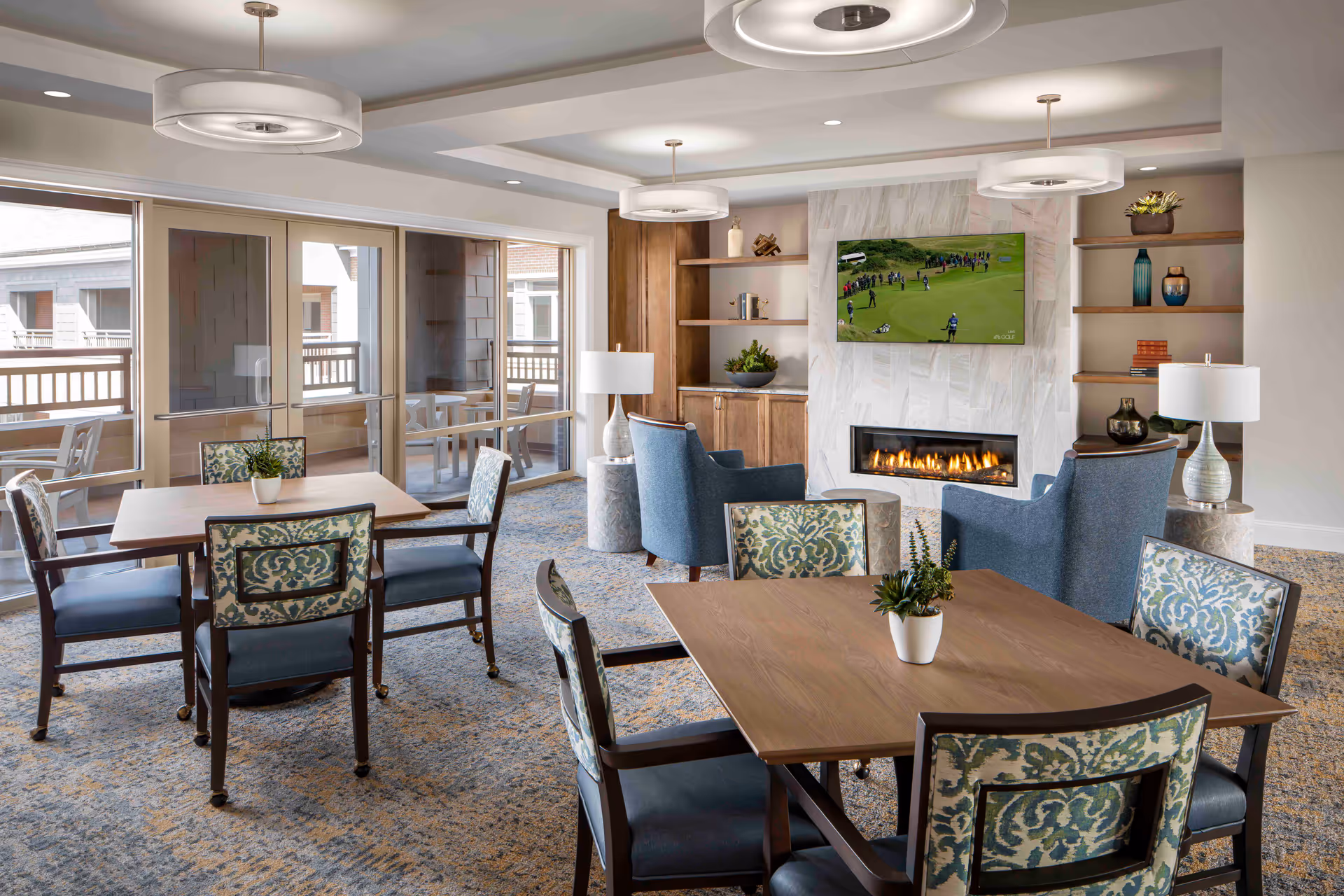 A cozy senior living common area with several wooden tables and upholstered chairs arranged on a patterned carpet. The room features a modern gas fireplace with a flat-screen TV mounted above it, displaying a golf game. Built-in wooden shelves flank the fireplace, decorated with plants, books, and vases. Large windows and glass doors lead to an outdoor balcony with additional seating. The ceiling has recessed lighting and three circular pendant lights.