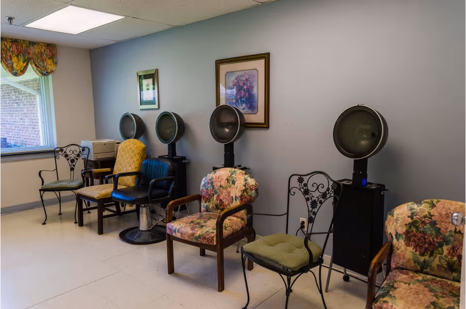 Small salon area with a row of hooded hair dryers and assorted chairs against a light blue wall.