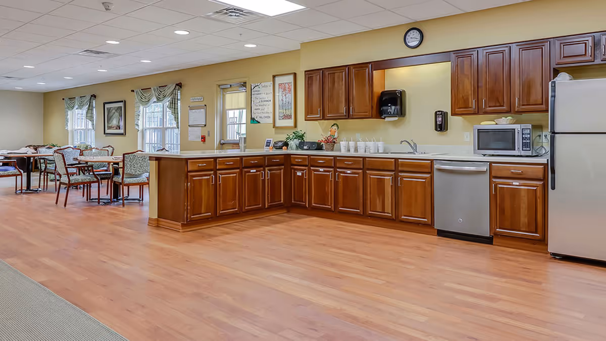 Communal dining area and kitchenette with wood cabinets, stainless refrigerator and dishwasher, and tables and chairs.