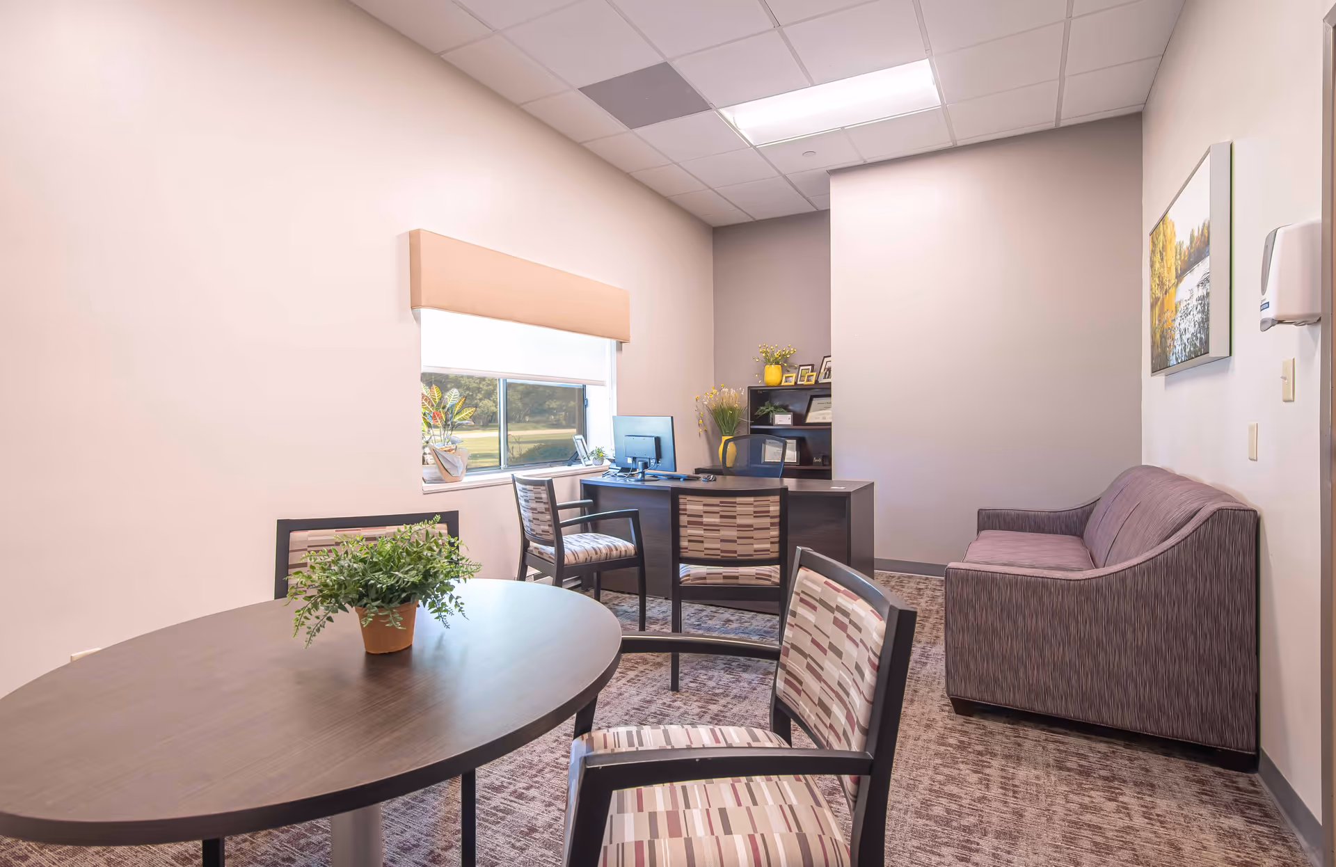 A small office room with a round table and three chairs with patterned upholstery. There is a potted plant on the table. Against the far wall is a desk with a computer and two chairs. A window with a beige valance lets in natural light. To the right is a purple upholstered loveseat and a wall-mounted soap dispenser. The room has light-colored walls and a carpeted floor.