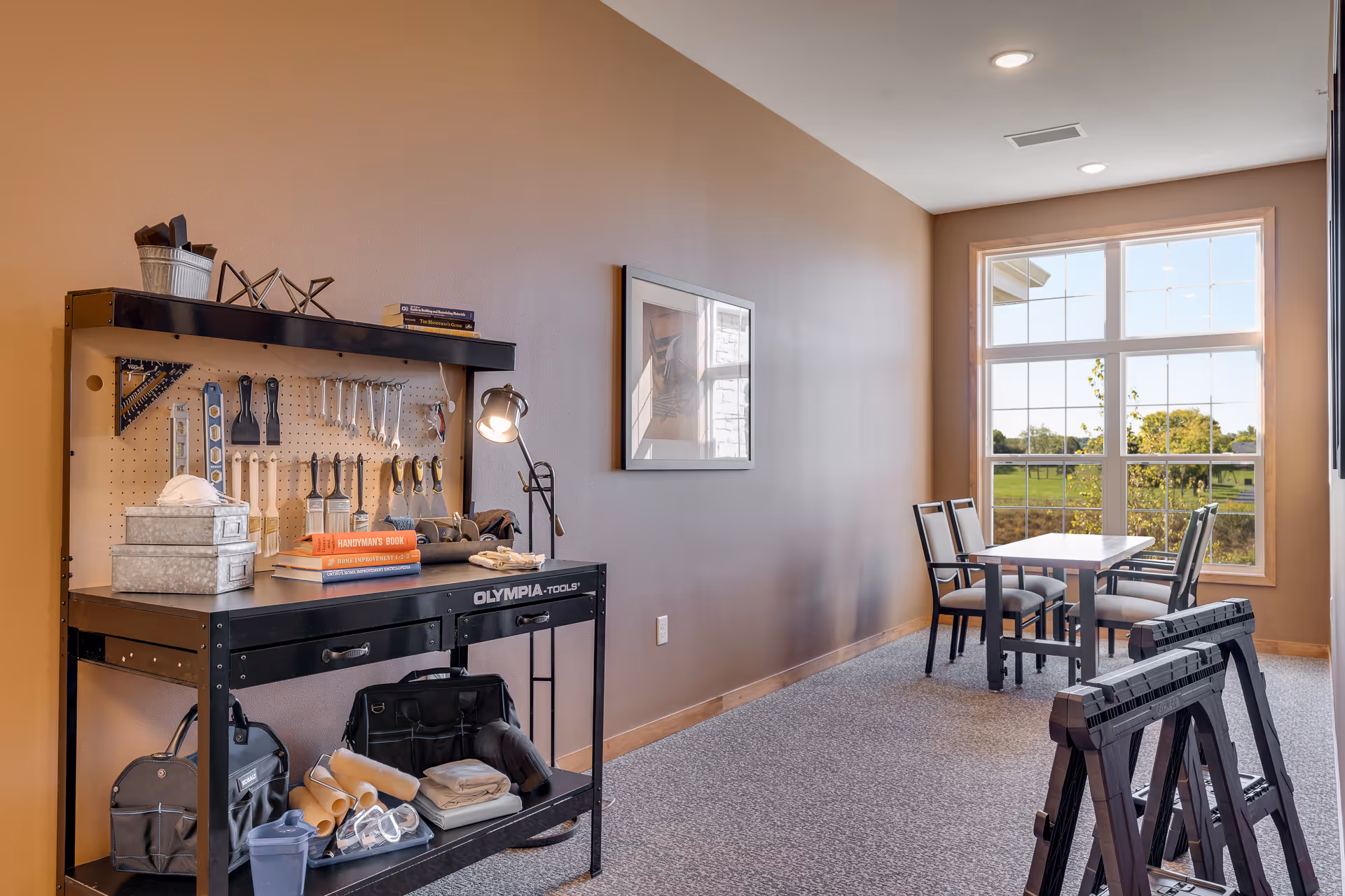 A well-lit room with a large window showing an outdoor view. On the left side, there is a black workbench with various tools hanging on a pegboard, books, and other supplies. On the right side near the window, there is a table with four chairs. The room has beige walls and carpeted flooring.