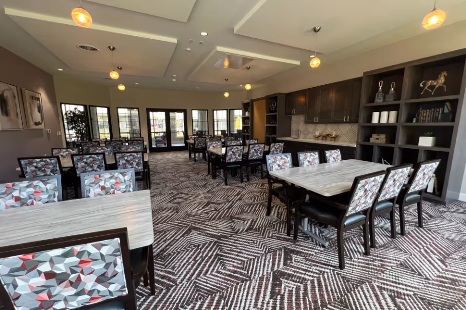 A spacious dining room with multiple tables and chairs featuring geometric patterned upholstery. The room has large windows and glass doors letting in natural light, pendant lights hanging from the ceiling, and built-in cabinetry with shelves displaying decorative items and books.