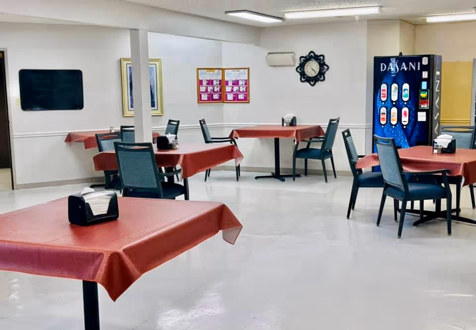 A clean dining area with several tables covered in red tablecloths and black chairs around them. A napkin holder is placed on each table. On the right side, there is a Dasani vending machine. The walls are white with a clock and framed pictures hanging. The floor is light-colored and shiny.