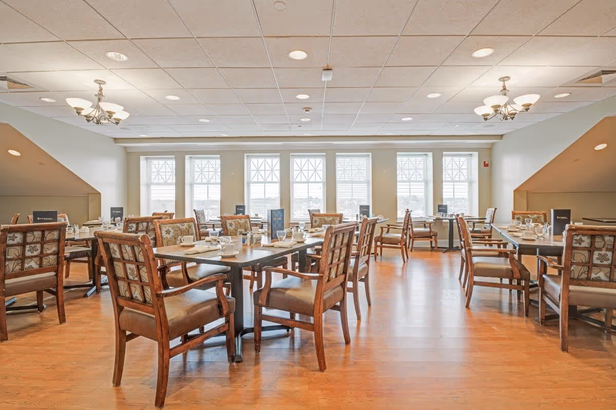 A bright dining room with wooden floors and multiple tables set with cups, plates, and menus. The room has large windows letting in natural light and wooden chairs with patterned cushions around the tables. Two chandeliers hang from the ceiling.