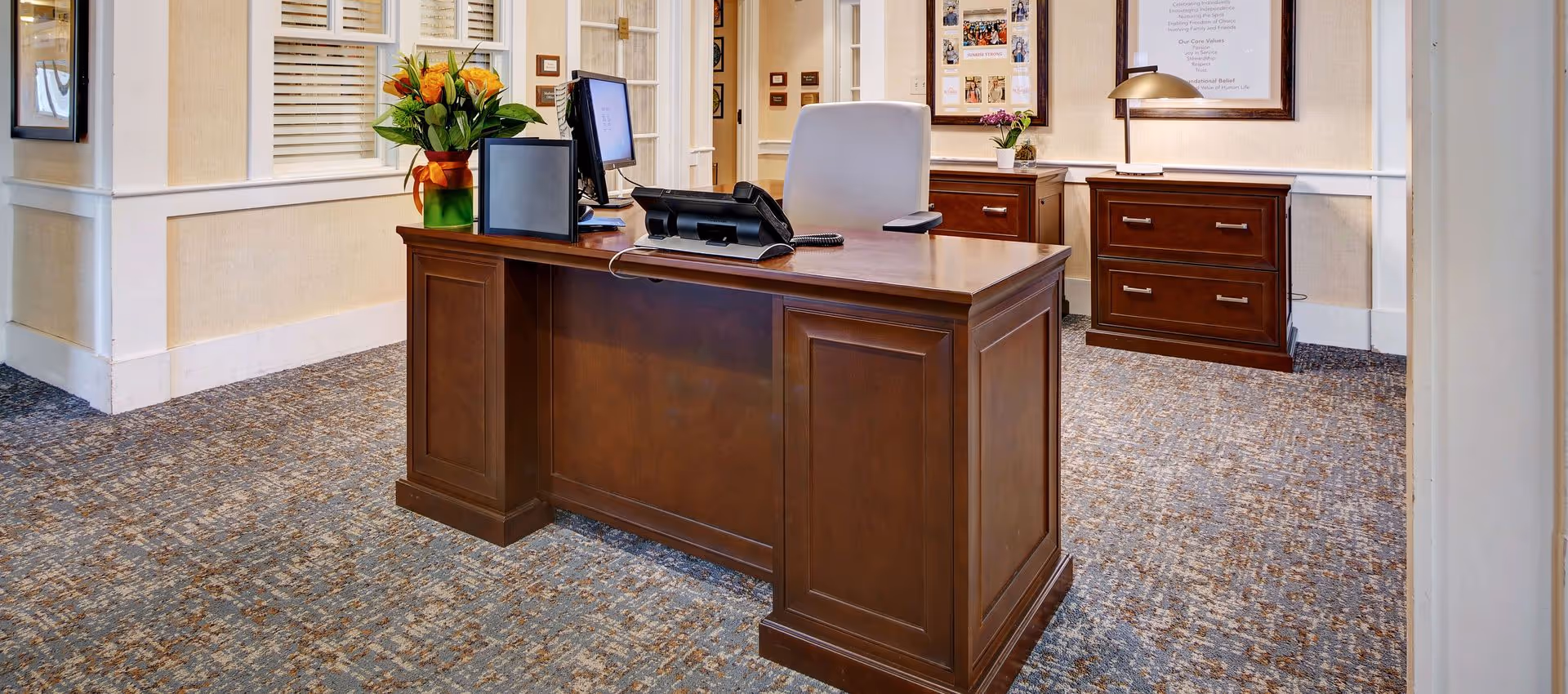 Reception desk area with a wooden desk, office chair, computer monitors, telephone, and a vase with orange flowers. The room has beige walls with white trim, framed pictures and documents on the walls, and a patterned carpeted floor.