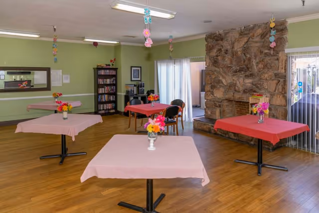 A common area with wooden flooring featuring four tables covered with pink and red tablecloths, each adorned with a vase of colorful flowers. The room has light green walls, a large stone fireplace, a bookshelf filled with books, and sliding glass doors letting in natural light. Hanging decorations are suspended from the ceiling.