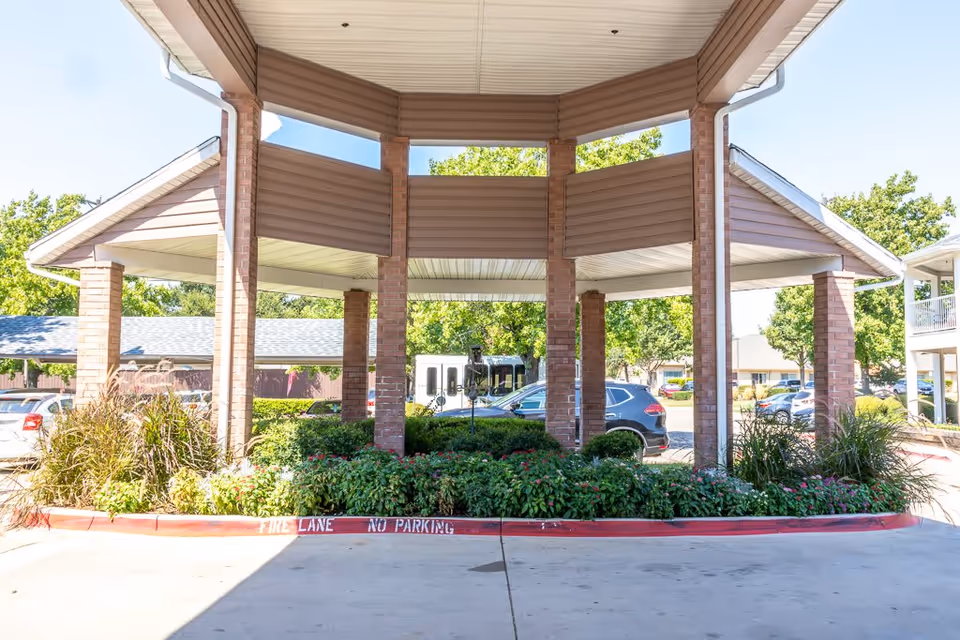 Covered entrance area with brick pillars and a landscaped garden bed in the center. Several parked cars and trees are visible in the background under a clear blue sky.