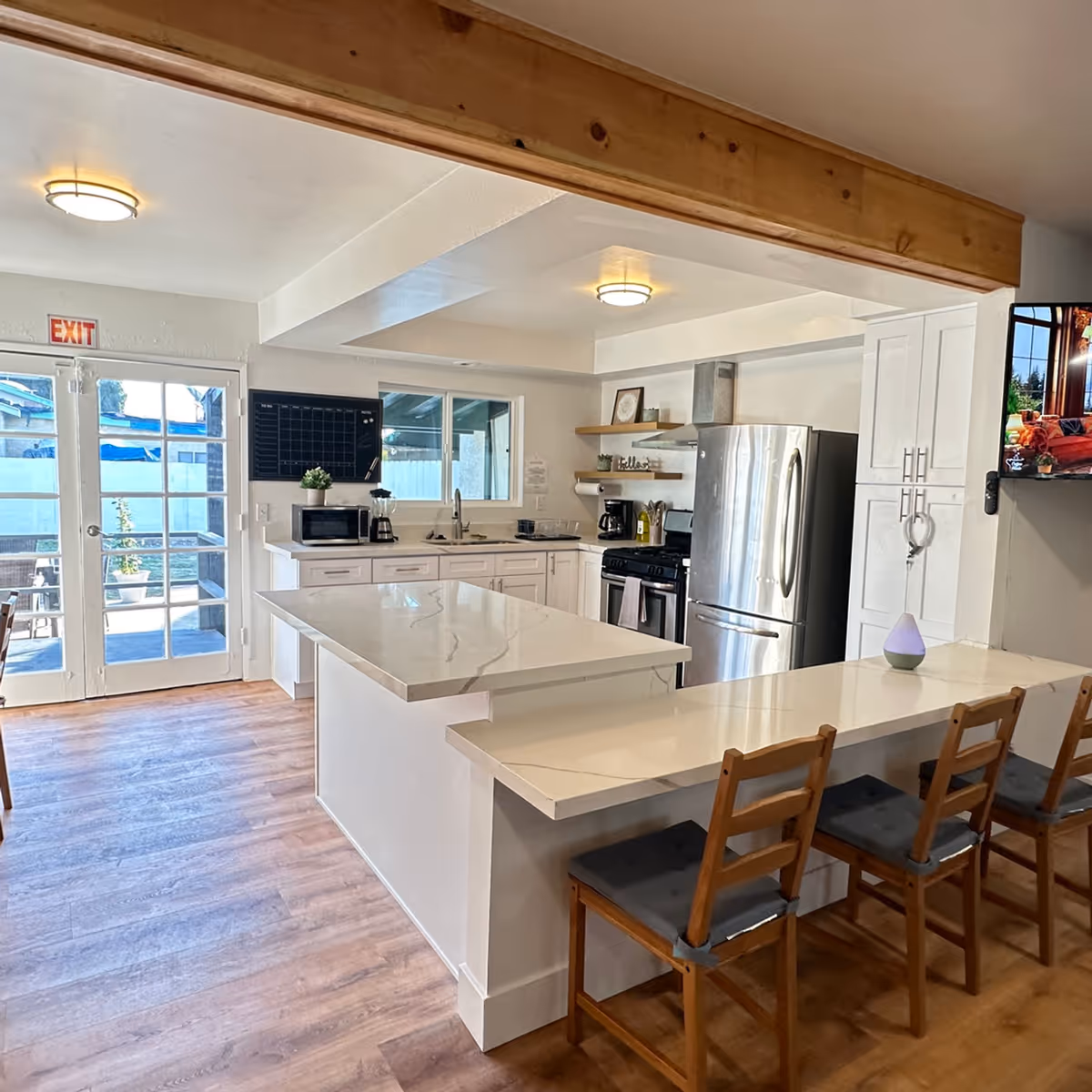 Modern kitchen area with white cabinets, a large island with a marble countertop, and a breakfast bar with wooden chairs. Stainless steel refrigerator and stove are visible. There is a window above the sink, a microwave and coffee maker on the counter, and a door leading outside. The floor is wooden and the ceiling has recessed lighting.