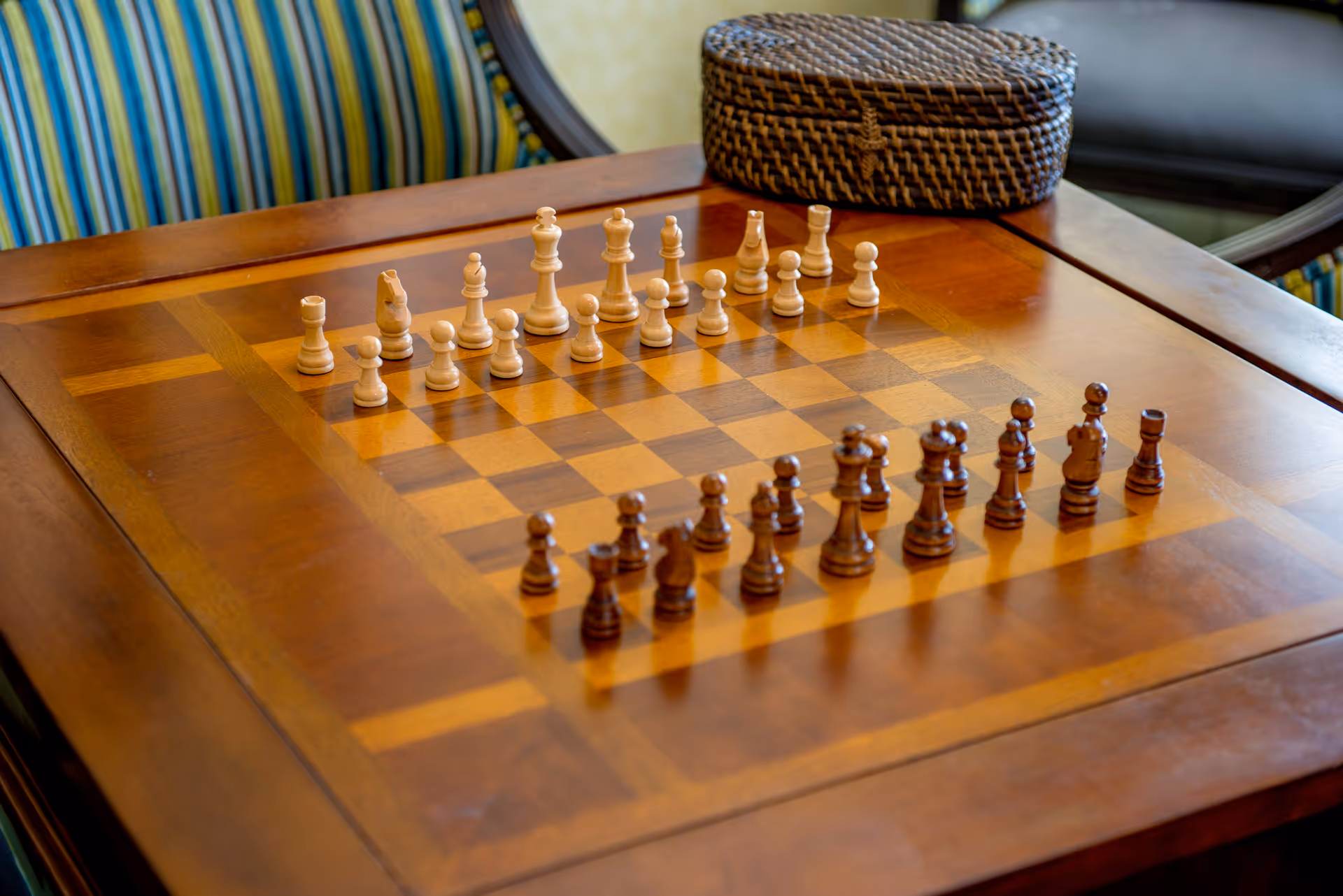 A wooden chessboard with chess pieces arranged in their starting positions on a polished wooden table. In the background, there is a striped upholstered chair and a woven basket on the table near a window.