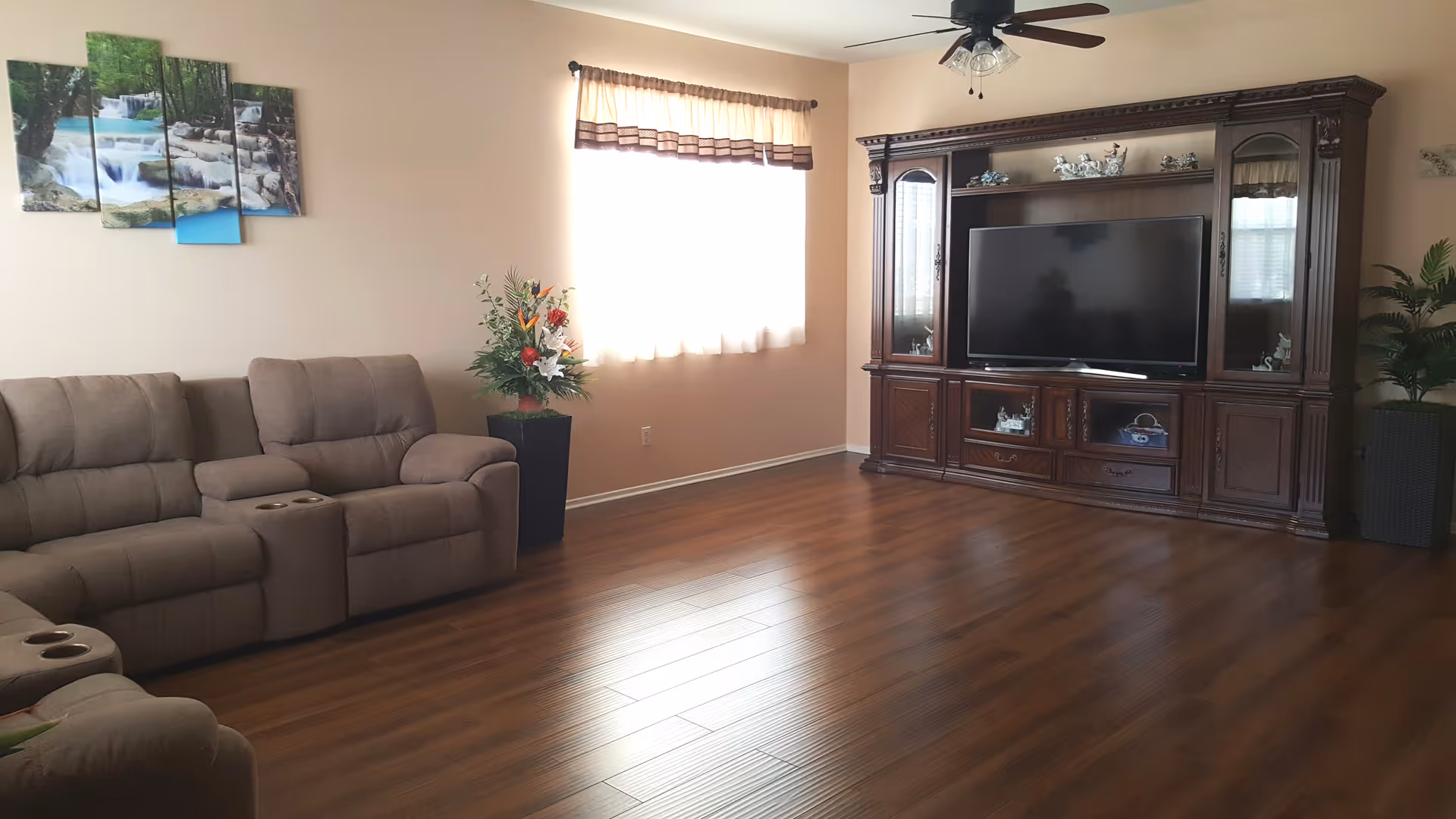 A living room with wooden flooring, a large brown sectional sofa with cup holders, a window with a curtain letting in natural light, a tall flower arrangement in a black vase, and a large dark wooden entertainment center with a flat-screen TV and decorative items.