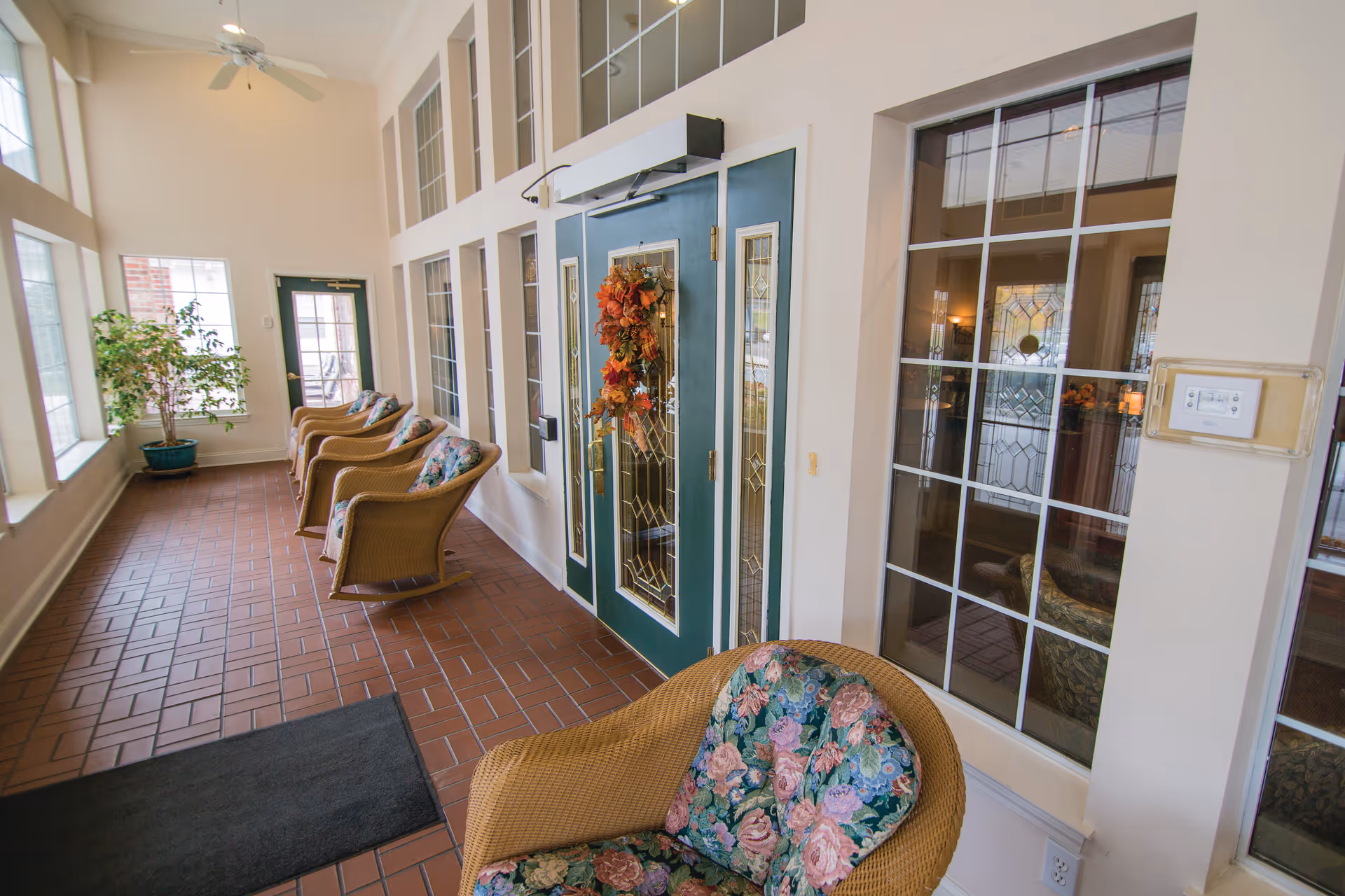 A bright indoor seating area with wicker chairs featuring floral cushions arranged along a wall with large windows. The floor is tiled with reddish-brown tiles, and there is a green door decorated with an autumn wreath. A potted plant is placed near the windows, and a ceiling fan is visible above.