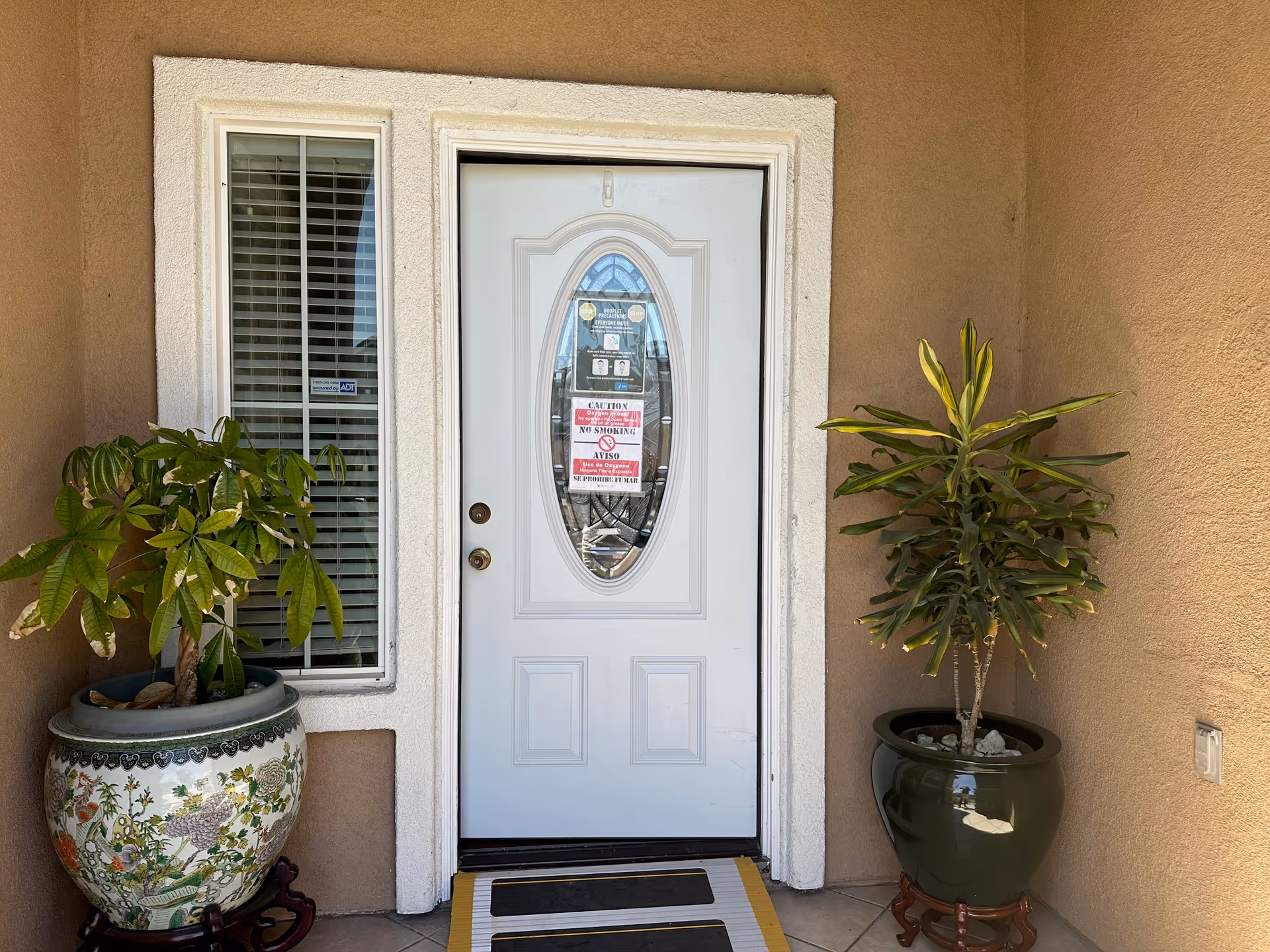 Front entrance door of a building with a white door featuring an oval glass window. There are two potted plants on either side of the door, one in a decorative ceramic pot and the other in a plain dark green pot. The door has multiple signs including a no smoking notice. The walls around the door are beige and there is a small window with blinds to the left of the door.