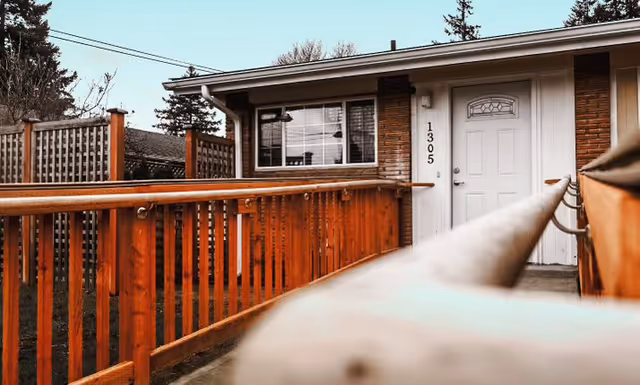 Exterior view of a single-story building with a white door and a window next to it. The building number 1305 is displayed vertically beside the door. A wooden handrail and fence lead up to the entrance, and trees are visible in the background under a clear sky.