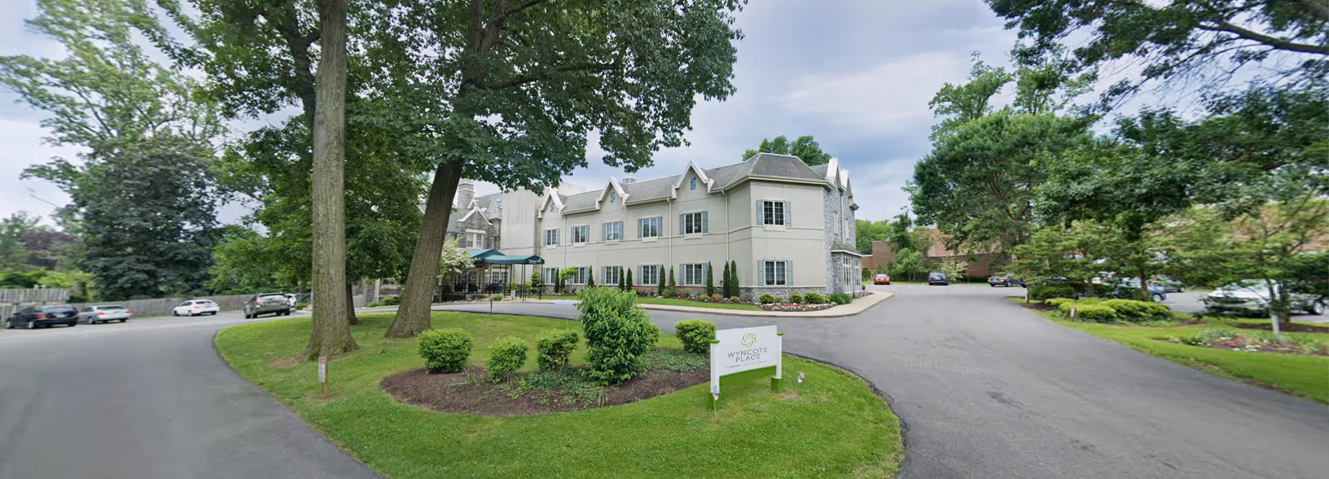 Exterior view of Wyncote Place, a multi-story senior living facility surrounded by trees and greenery. The building has a light-colored facade with multiple windows and a covered entrance. There is a driveway and parking area with several cars parked. A sign with the facility's name is visible on a grassy area near the driveway.