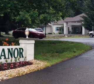 Entrance driveway to a senior living facility with a white brick sign partially visible with the word 'MANOR' in green letters, a landscaped area with flowers and rocks, a red car parked near the entrance, and a single-story building with a covered entryway in the background surrounded by trees.