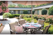 Outdoor patio area with a round table set for four, featuring plates, glasses, and napkins. The table is shaded by a large umbrella. Surrounding the patio are well-maintained bushes, flowering plants, and a building with windows in the background.