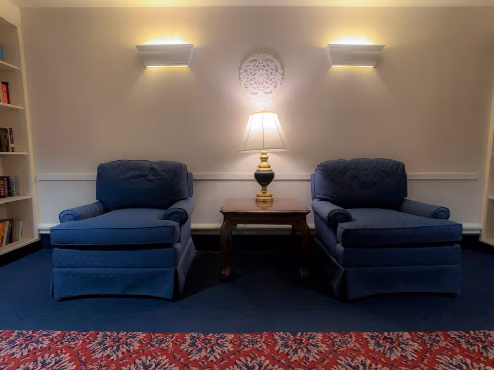 Two blue upholstered armchairs with a wooden side table and a lamp between them, positioned against a beige wall with two wall sconces and a decorative wall piece above the lamp. A bookshelf is partially visible on the left side and a patterned red carpet is on the floor.