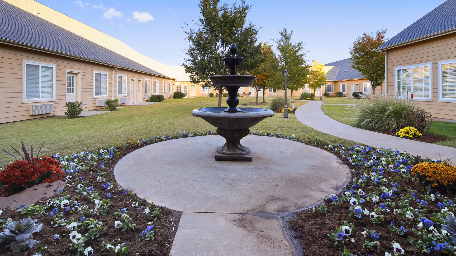 Outdoor courtyard area of a senior living facility with a central black water fountain surrounded by flower beds with purple and white flowers. The courtyard is bordered by beige buildings with white-framed windows and doors, and there are trees and a curved concrete pathway leading through the grassy area.