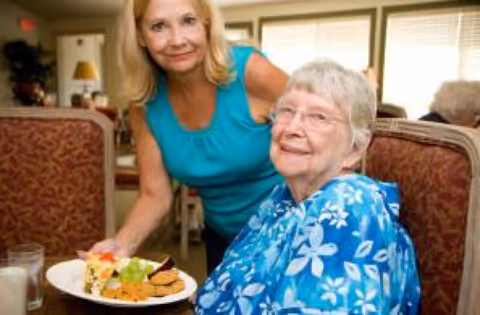An elderly woman seated at a dining table with a plate of food in front of her, accompanied by a younger woman standing beside her, both smiling in a well-lit dining room setting.