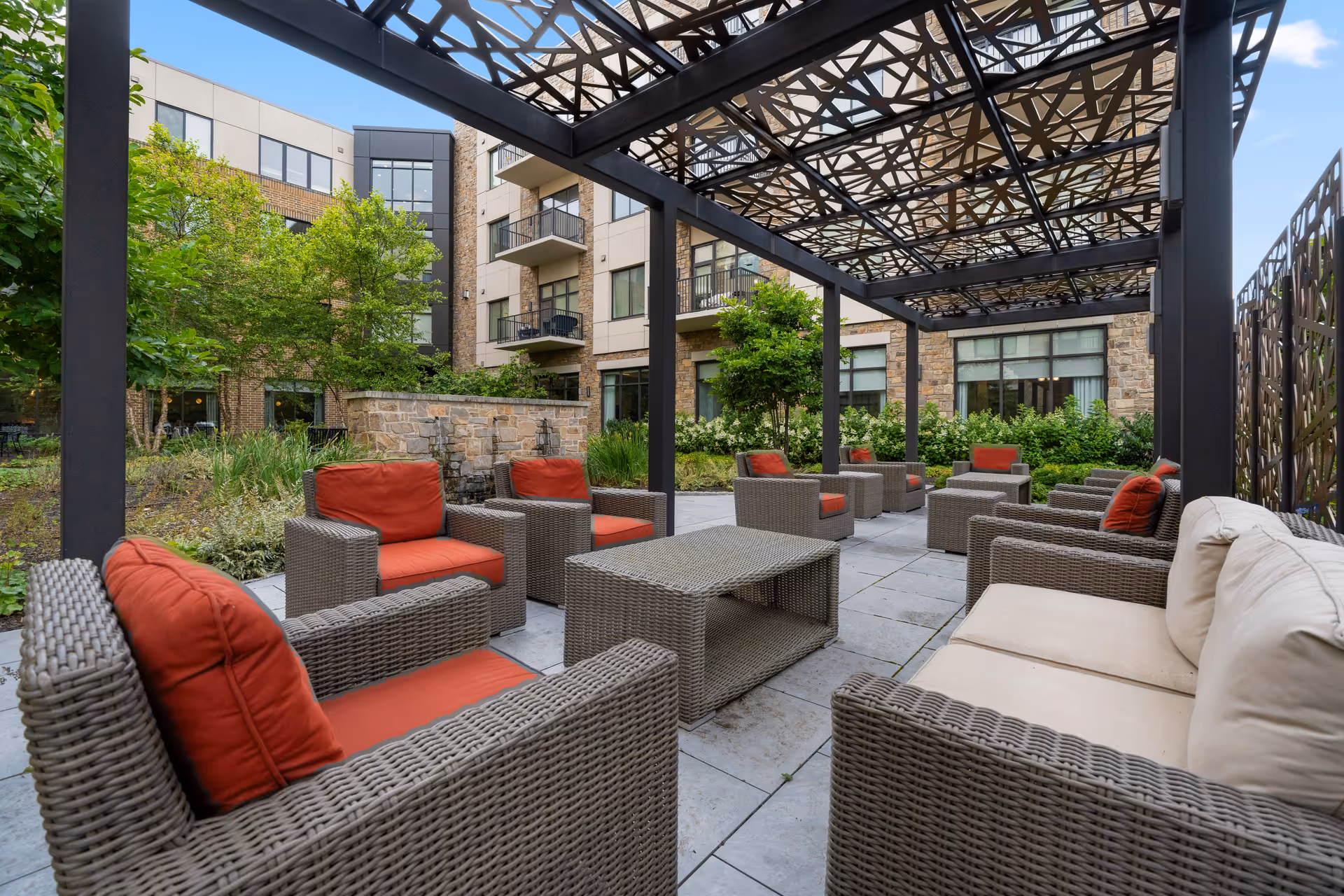 Covered outdoor courtyard with wicker lounge seating and red cushions in front of a multi-story building.
