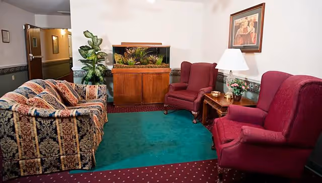 A cozy sitting area in an assisted living facility featuring a patterned sofa with cushions, two red upholstered armchairs, a wooden side table with a lamp and a small plant, a large fish tank on a wooden cabinet, and a framed artwork on the wall. The floor has a green carpet bordered by a red carpet with white dots, and there is a potted plant near the hallway entrance.