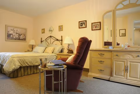 A cozy bedroom with a large bed featuring a metal headboard, two bedside tables with lamps, a comfortable maroon recliner chair, a small round side table with books and a glass, and a wooden dresser with a large mirror. The walls are decorated with framed pictures and the room has a warm, inviting atmosphere.